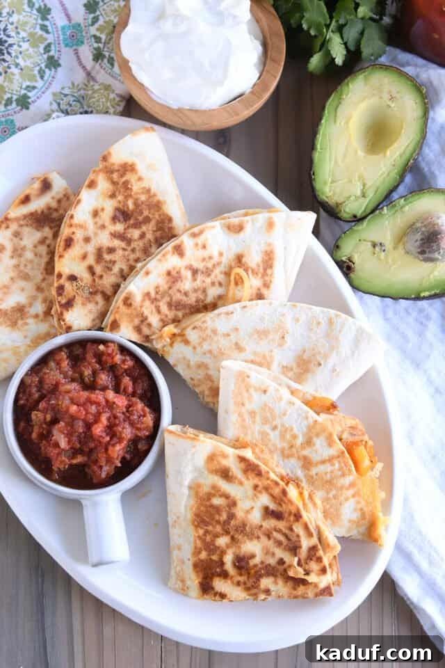 Irresistible Chicken Quesadillas 7 A white platter showcasing six perfectly cut chicken quesadilla triangles, beautifully arranged with bowls of vibrant salsa, sliced avocado, and creamy sour cream for serving.