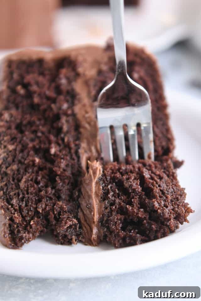 Next-Level Chocolate Cake Perfection 3 A fork gently lifts a perfect piece of frosted chocolate cake from a white plate, showcasing its soft, moist crumb and rich appearance.