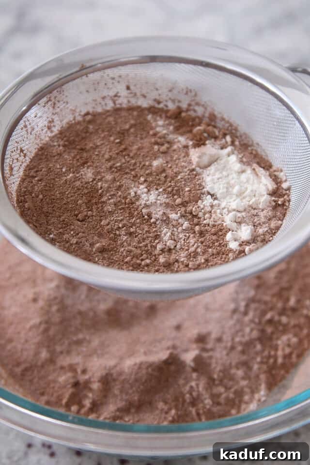 Next-Level Chocolate Cake Perfection 4 A close-up shot of dry ingredients – cocoa powder, flour, and sugar – being sifted through a mesh strainer into a large glass bowl, illustrating a crucial step for a smooth cake batter.