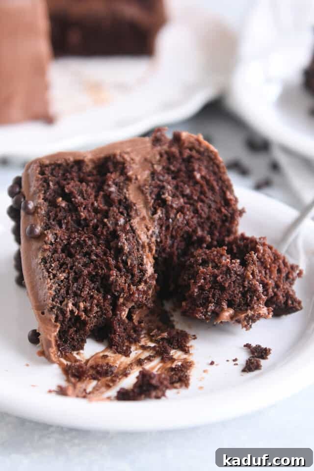 Next-Level Chocolate Cake Perfection 9 A perfectly portioned slice of chocolate cake, generously frosted with smooth chocolate, rests invitingly on a white plate, ready to be enjoyed with a fork.