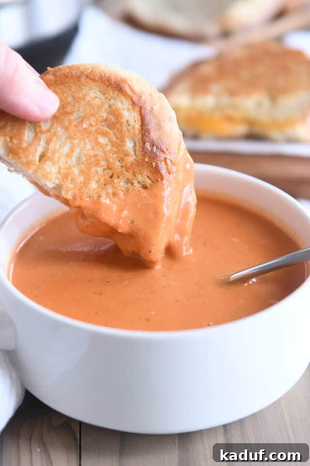 Velvety Instant Pot Tomato Basil Delight 6 A hand dipping half of a golden grilled cheese sandwich on sourdough bread into a rustic white bowl of creamy tomato basil soup.