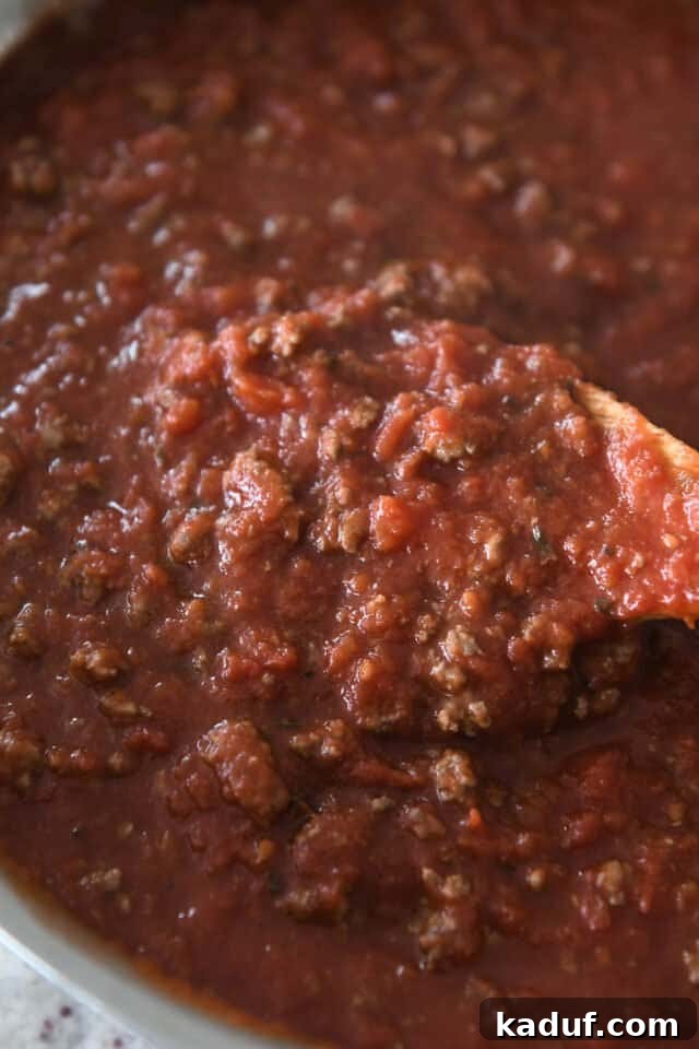 Your Go-To Cheesy Meal Prep Pasta Bake 3 A wooden spoon stirring vibrant red meat sauce with cooked ground beef in a shining stainless steel pan, showcasing the rich texture.