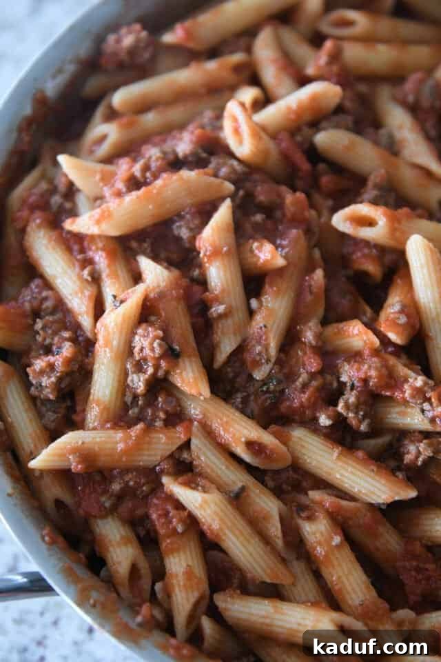 Your Go-To Cheesy Meal Prep Pasta Bake 4 Penne noodles generously coated in rich red meat sauce within a large stainless steel pan, ready for the next layer of cheesy goodness.