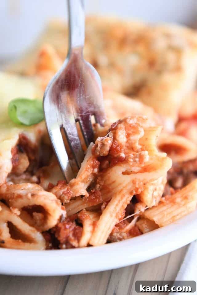 Your Go-To Cheesy Meal Prep Pasta Bake 7 A fork taking a delicious bite of penne noodles coated in red sauce and cheese on a white plate, highlighting the inviting texture.