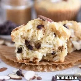 One almond coconut chocolate chip muffin broken in half on brown paper liner.