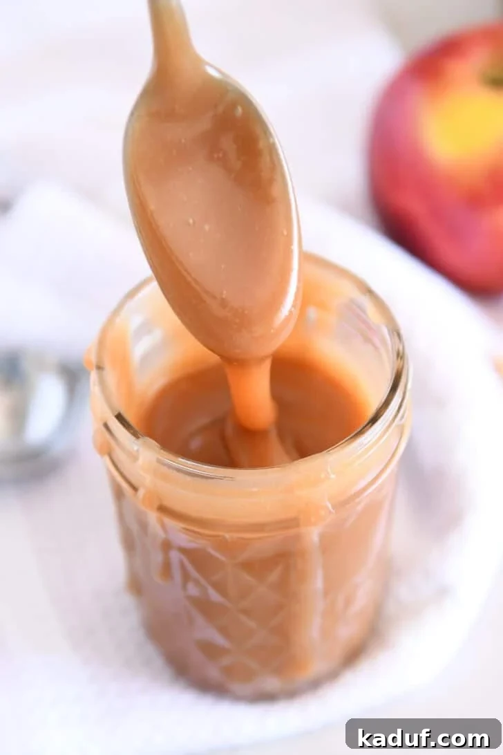 Spoon drizzling caramel sauce into glass jar with apple in the background.