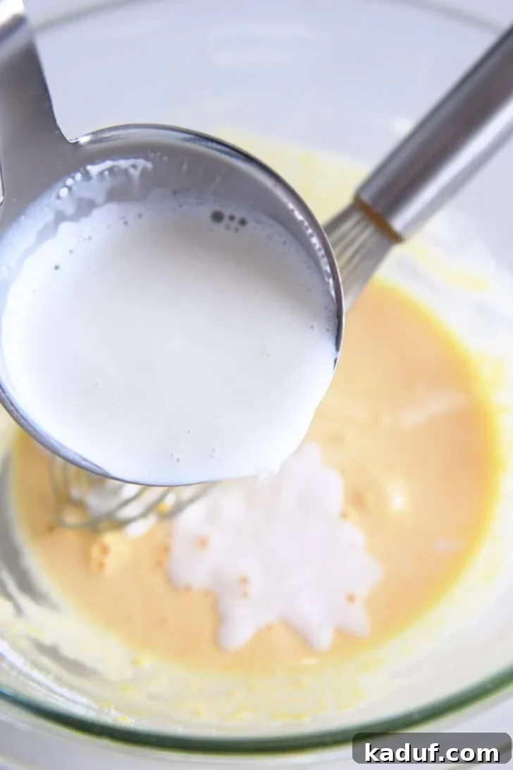 Homemade Banana Pudding from Scratch: Creamy Classic Recipe 5 Pouring ladle of hot cream into tempered egg yolks and sugar in glass bowl.