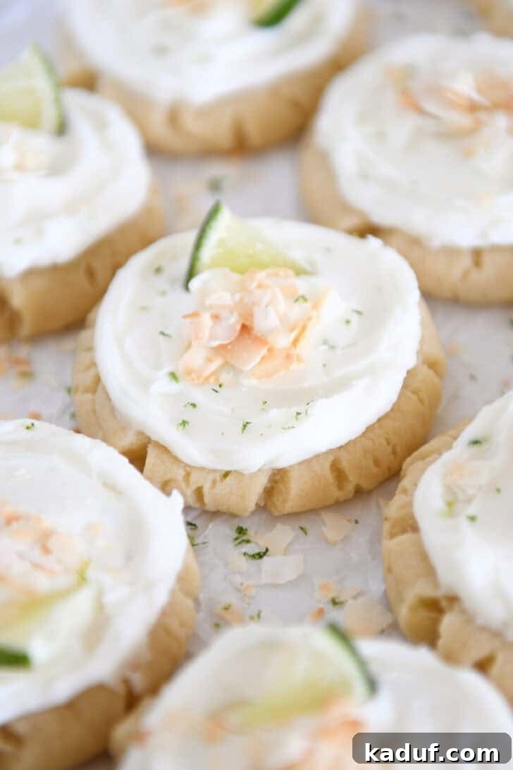 Lots of frosted coconut lime sugar cookies topped with toasted coconut and a fresh lime wedge on white parchment paper.