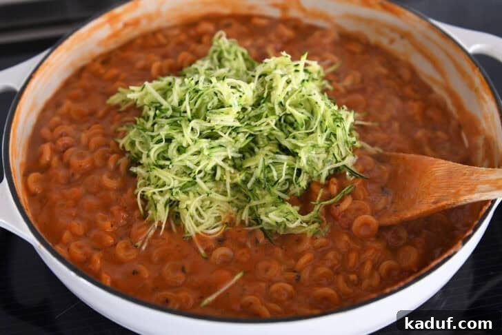 One-Pot Saucy Zucchini Pasta for Quick Weeknight Meals 5 Adding shredded zucchini to cooked pasta in red sauce with shredded zucchini in deep skillet.